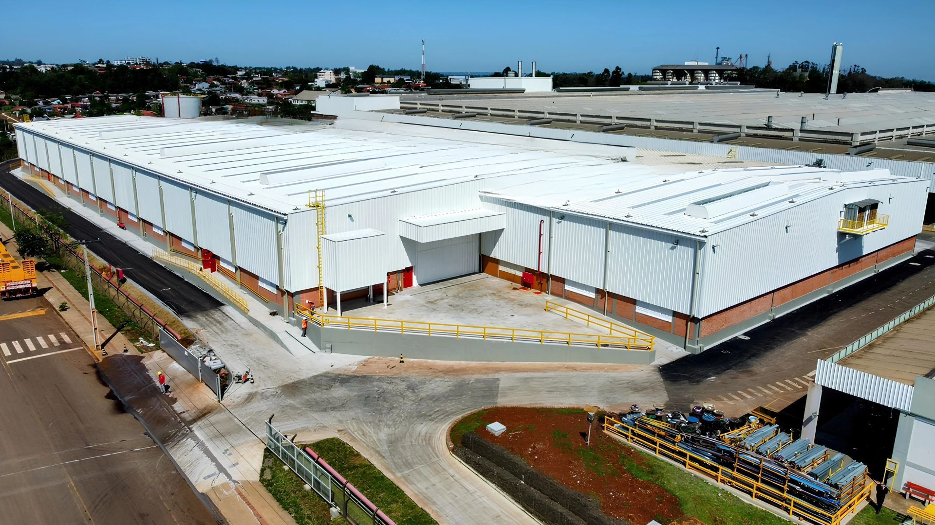 Grande complexo de armazéns industriais brancos em construção, cercado por estradas e terraplenagens.