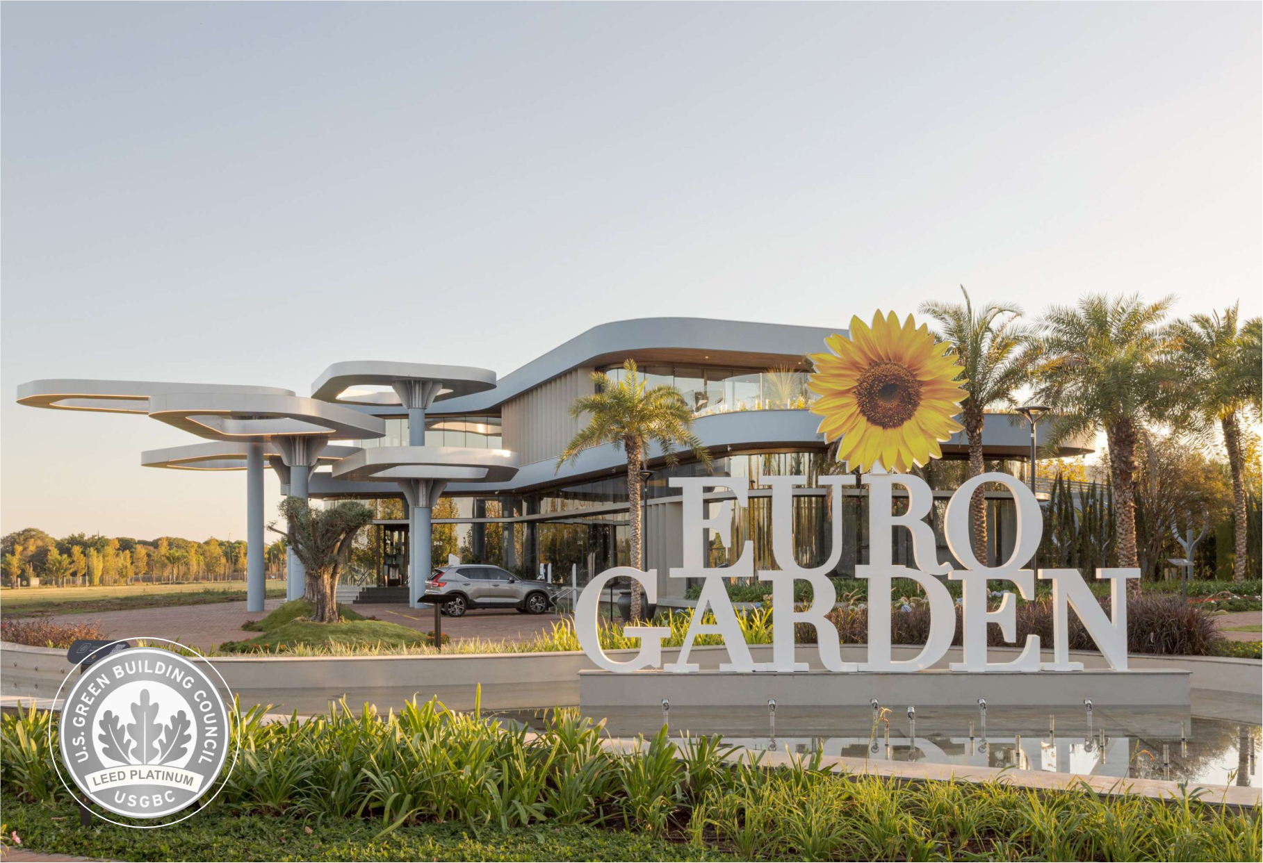 Placa de entrada do Modern Euro Garden com escultura de girassol, palmeiras e jardins paisagísticos ao pôr do sol.