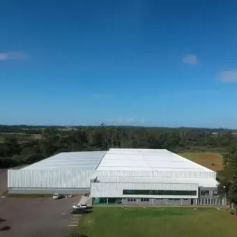 Vista aérea de um grande edifício industrial branco com céu azul e árvores ao fundo.