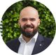 Homem careca sorridente, com barba, vestindo terno escuro e camisa branca, em pé diante de um fundo verdejante.