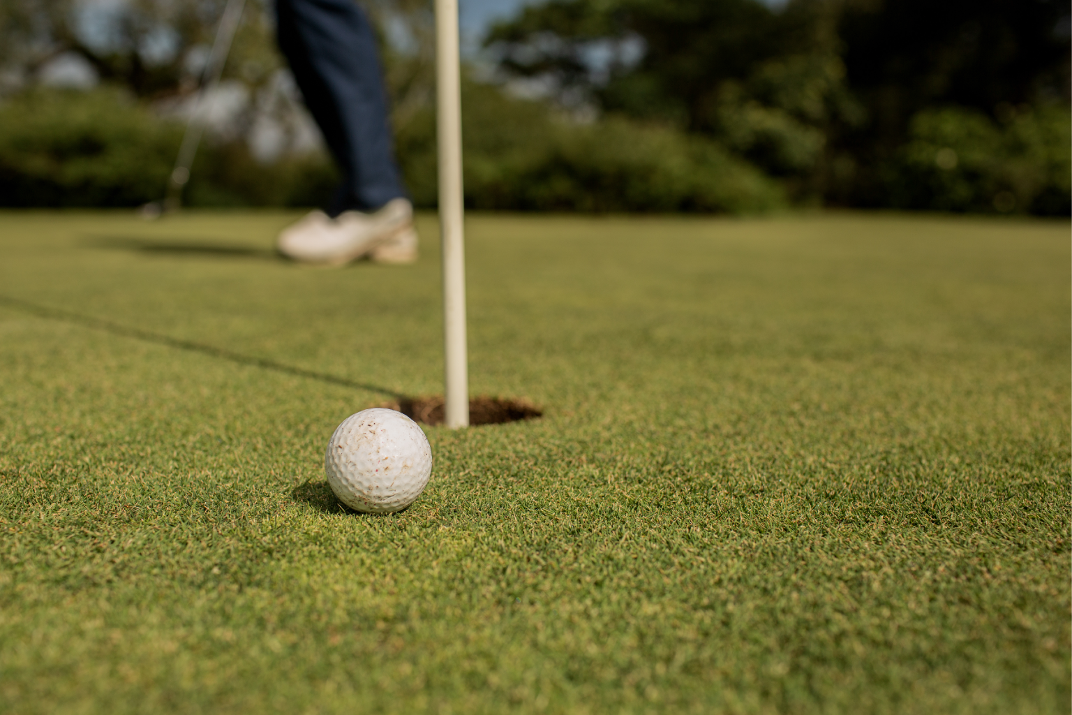 Bola de golfe perto de um buraco em um green, com um jogador de golfe ao fundo.