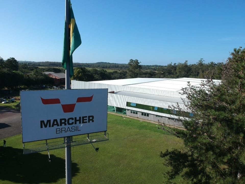 Placa com os dizeres “MARCHE BRASIL” ao lado de uma grande tenda branca para eventos e uma bandeira em uma encosta gramada.
