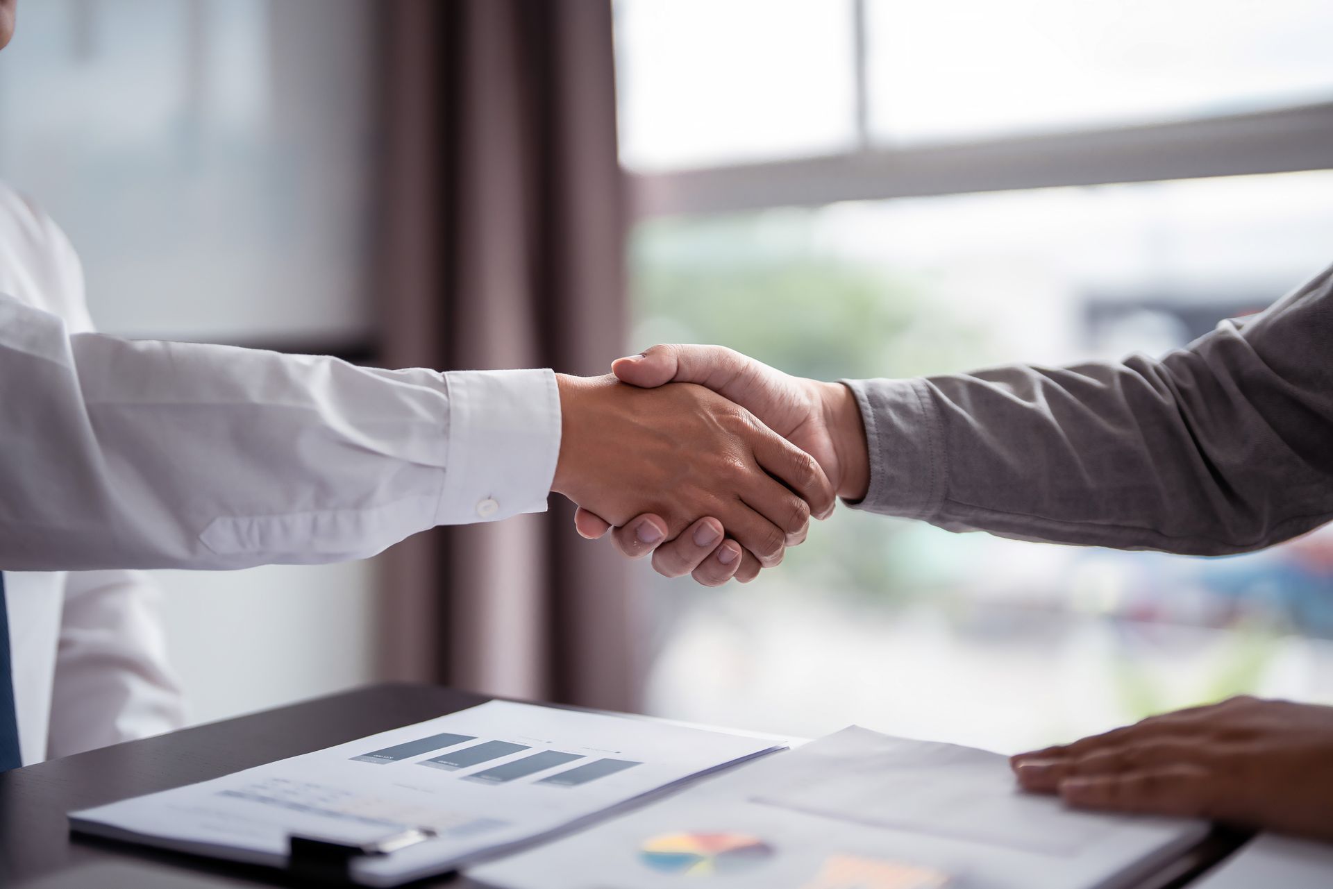 Two people shake hands across a desk in a bright office.