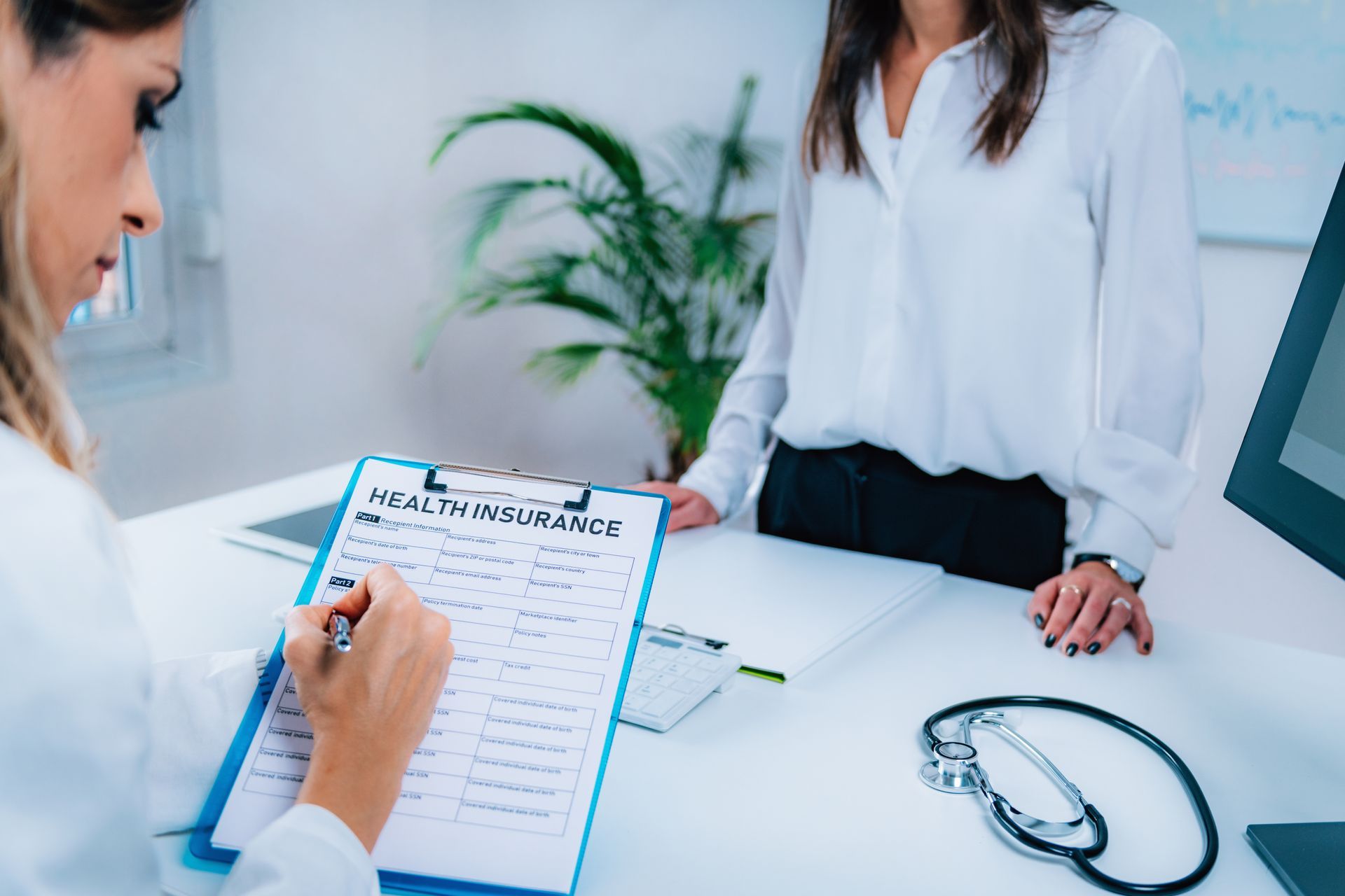 Doctor filling out a medical form at a desk while another clinician stands nearby with a stethoscope.