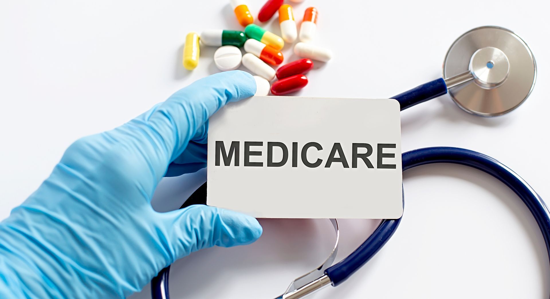 Gloved hand holding “MEDICARE” card beside pills and a stethoscope on a white background