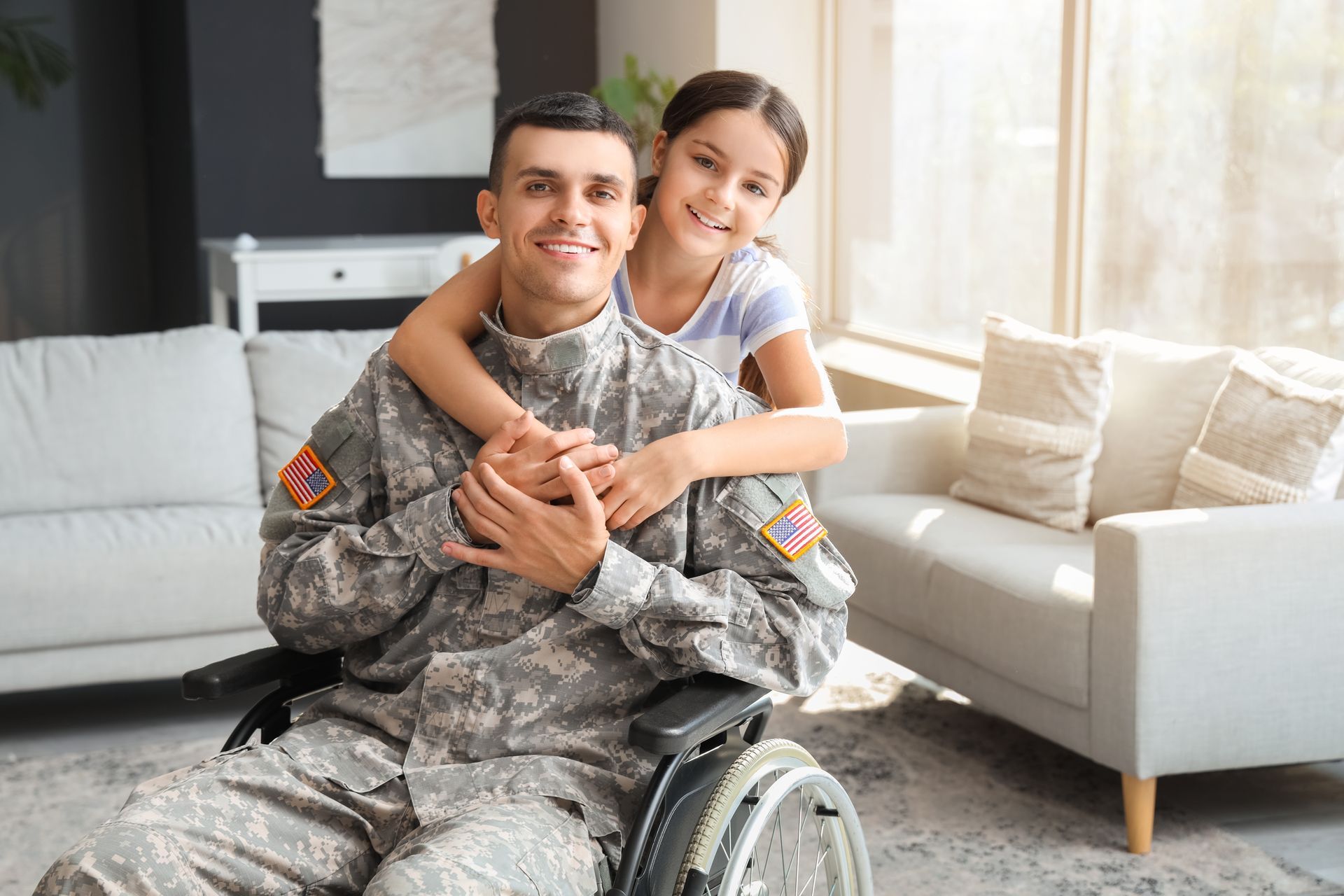 Smiling person in camouflage military uniform and wheelchair with child hugging them on a bright living room couch