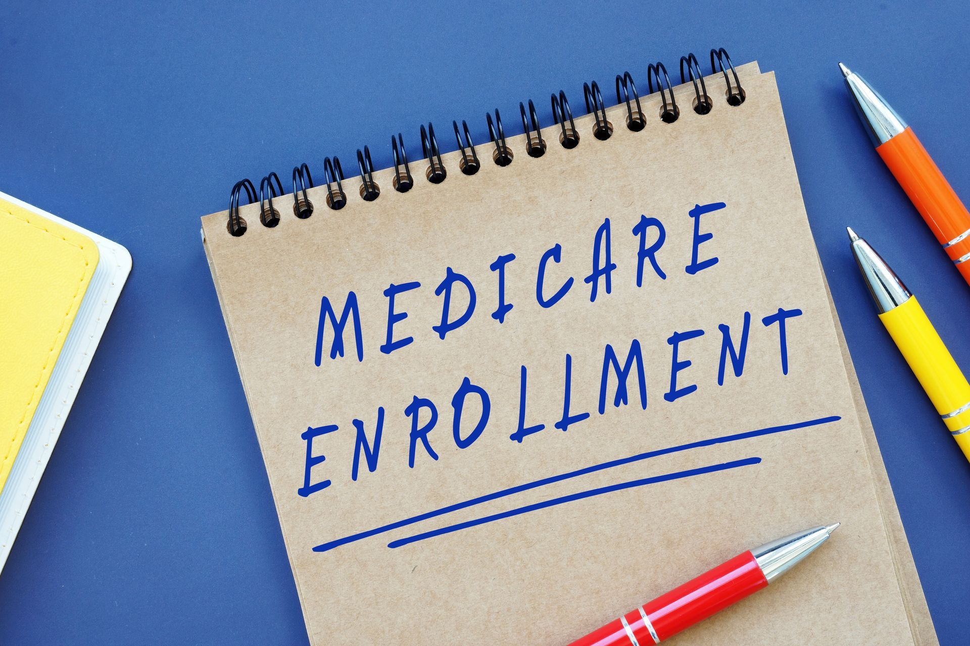 Notebook with “Medicare Enrollment” written on it, surrounded by colorful pens on a blue desk.