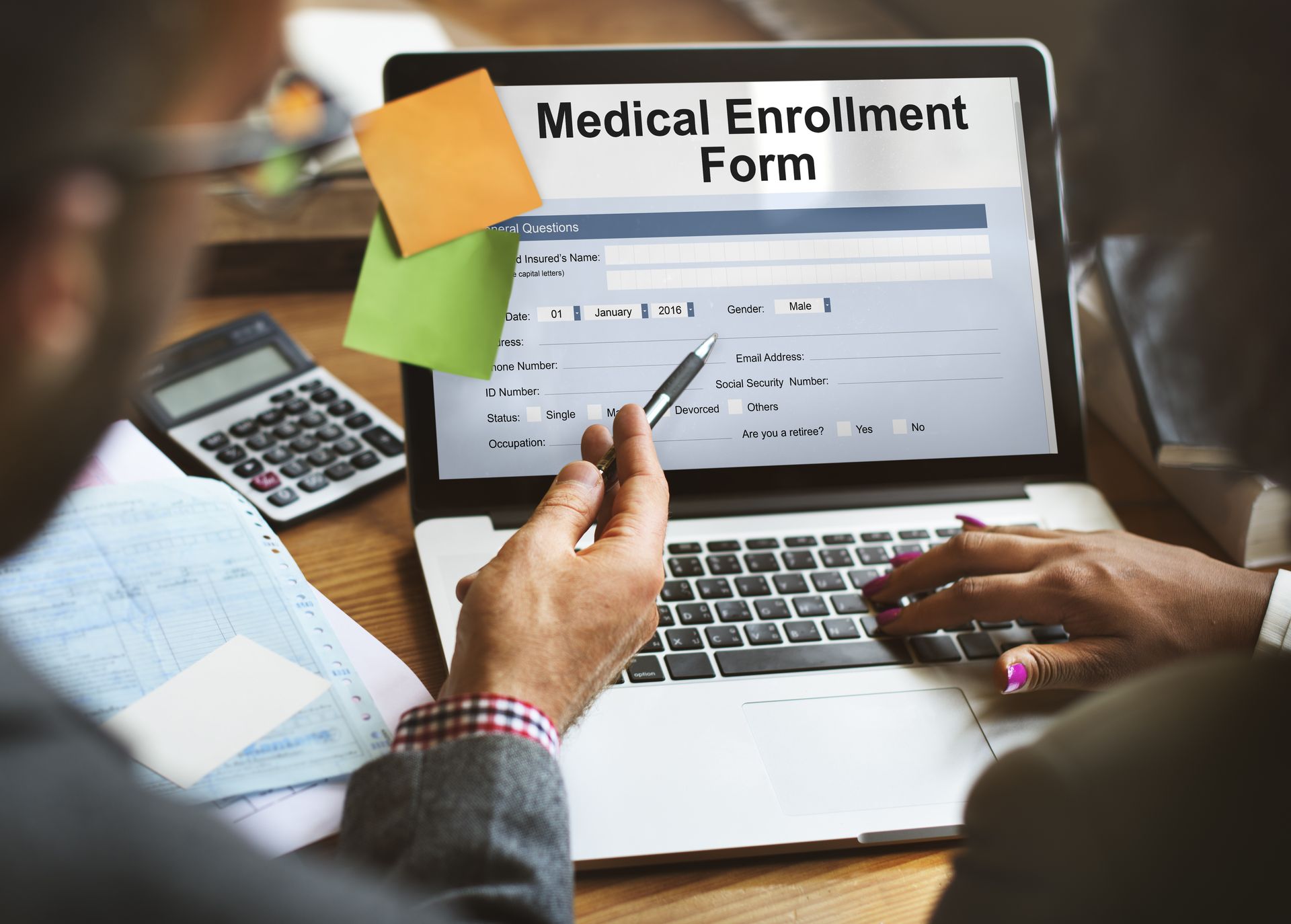 People filling out a medical enrollment form on a laptop, with calculator and sticky notes nearby