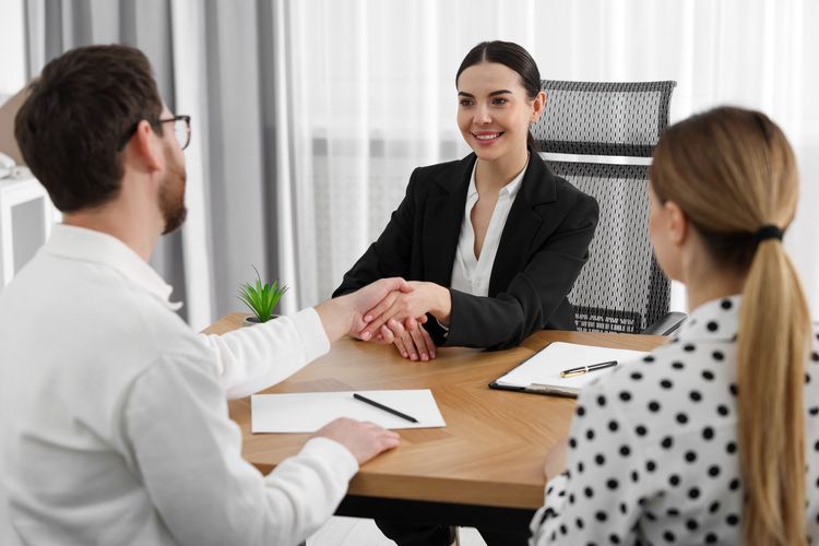 Three people in a meeting shake hands across a wooden table in a bright office.