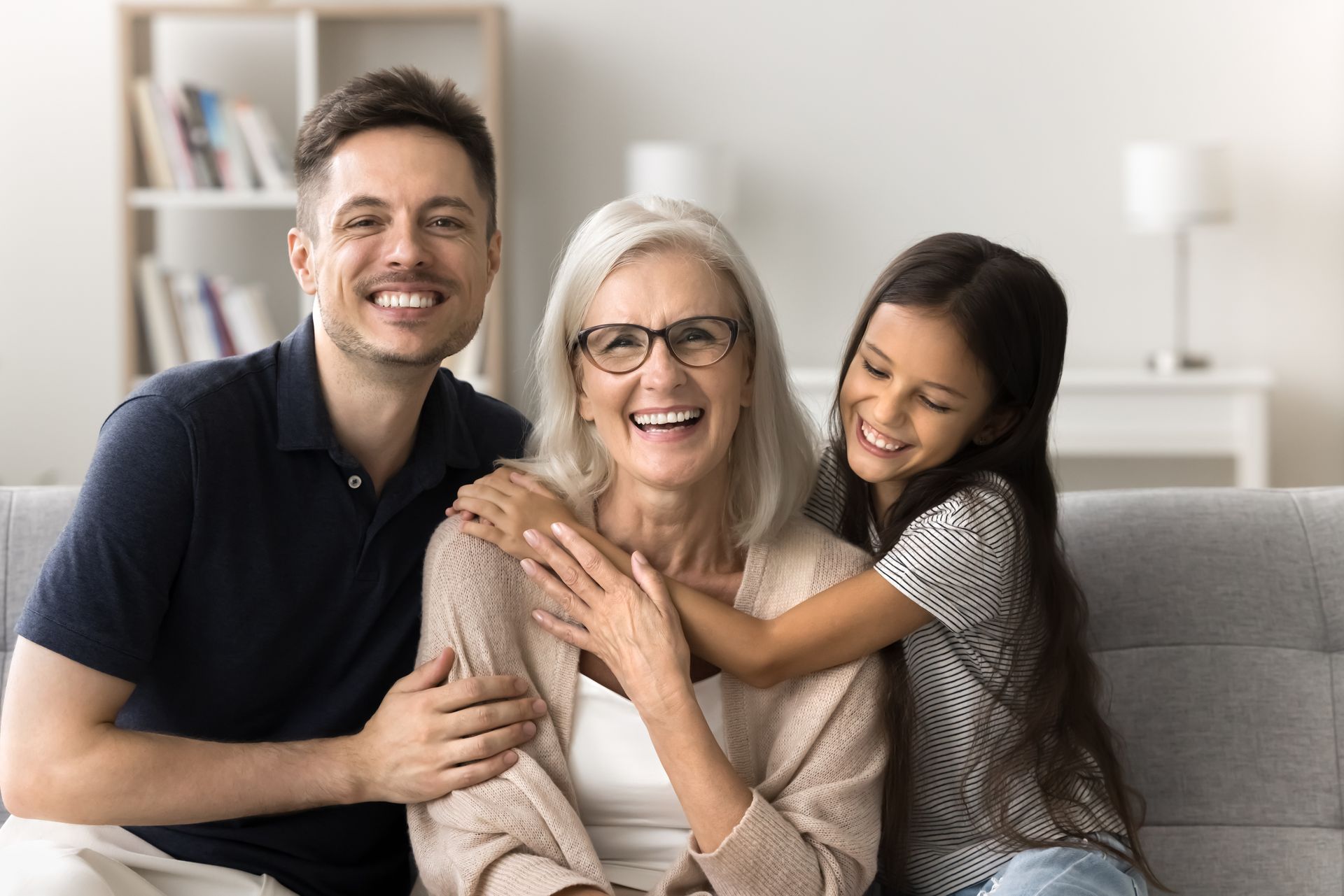 Three people smiling on a couch, with one woman hugging an older woman in a bright living room.