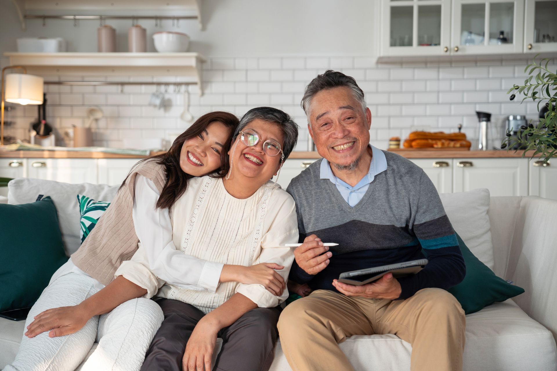 Three smiling people sit on a white couch in a bright kitchen, sharing a tablet