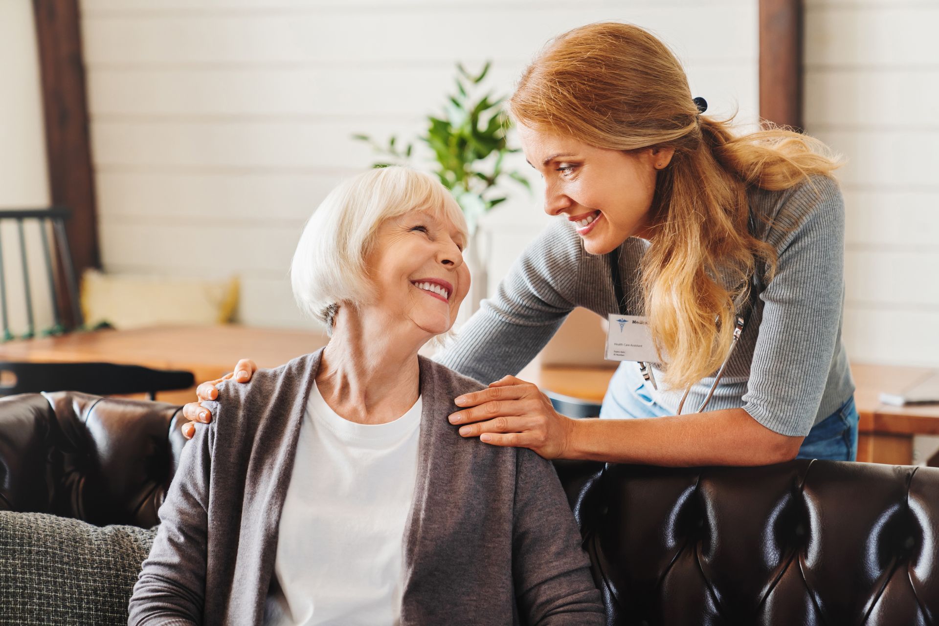 Woman smiling with caregiver on a dark leather couch in a bright living room