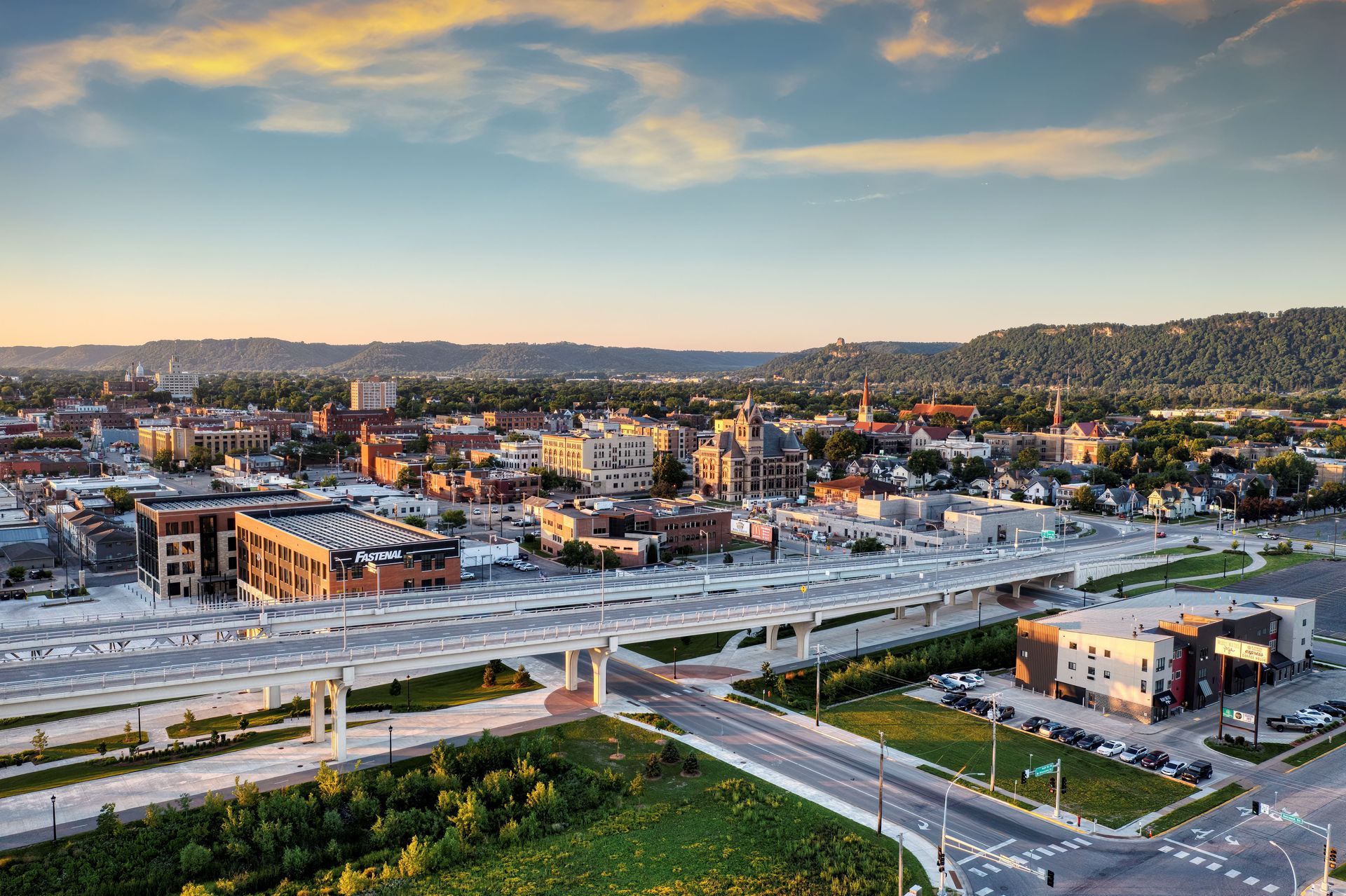 Aerial view of a city at sunset with buildings, roads, and hills under a pastel sky