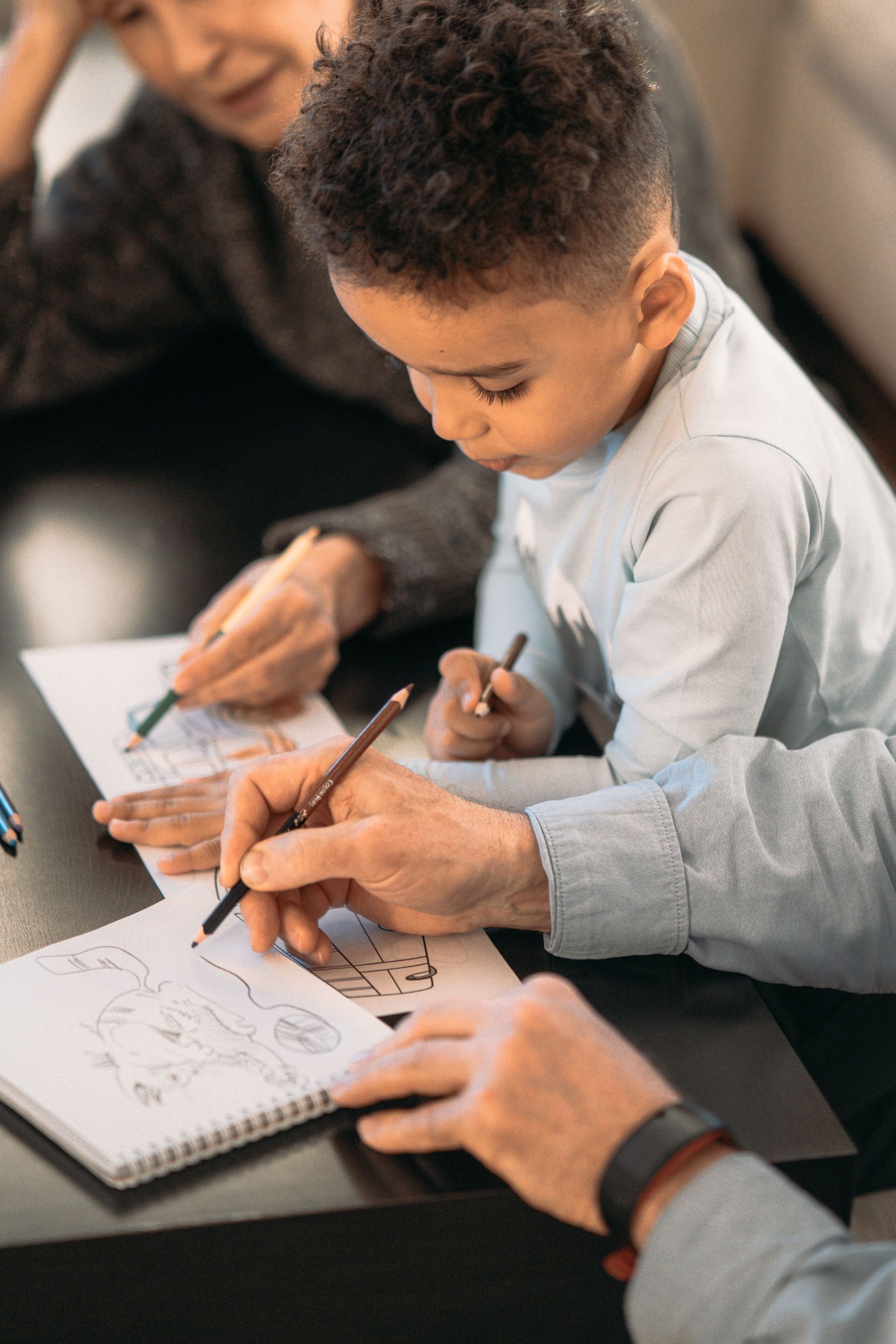 Two people and a child writing in notebooks at a table, hands close-up in a classroom or meeting setting