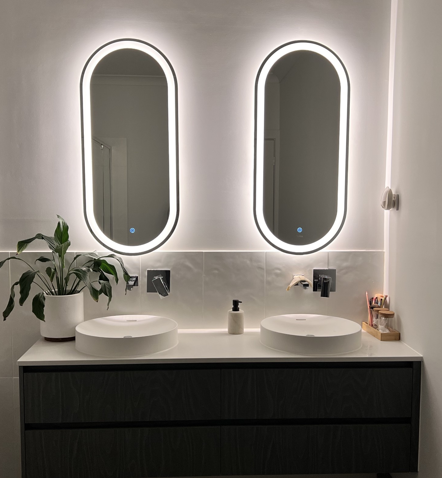 Modern bathroom vanity with two illuminated oval mirrors, double sinks, and a potted plant on the counter. By Plan Interior Architecture