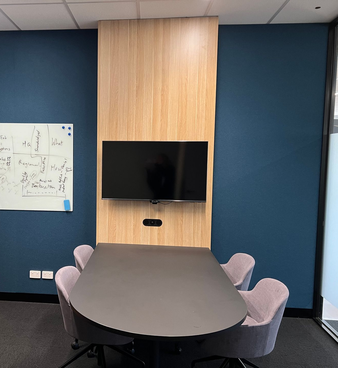 Small meeting room with oval table, upholstered chairs, wall-mounted TV, and whiteboard on blue walls. By Plan Interior Architecture