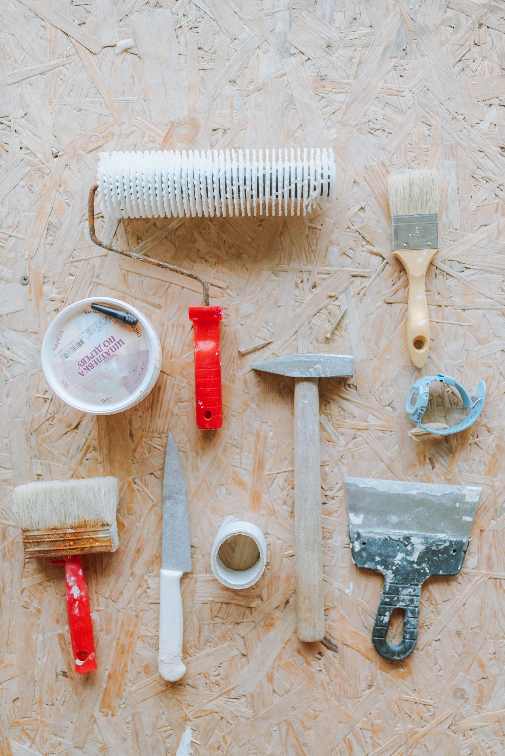 Assorted paint and wallpaper tools arranged on a light surface, including brushes, roller, scraper, and knife