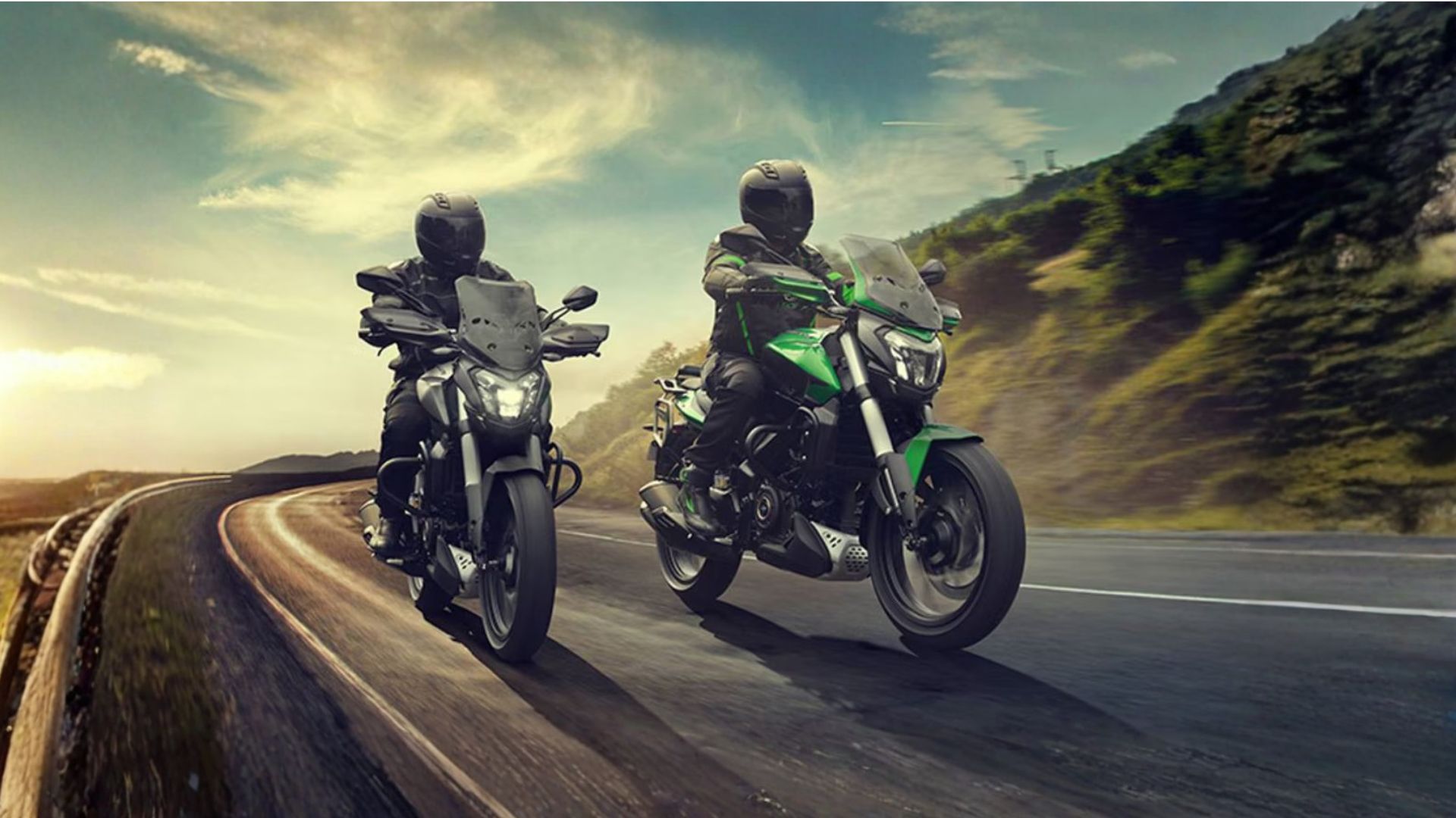 Two motorcyclists riding on a curving road through a mountainous landscape at sunset