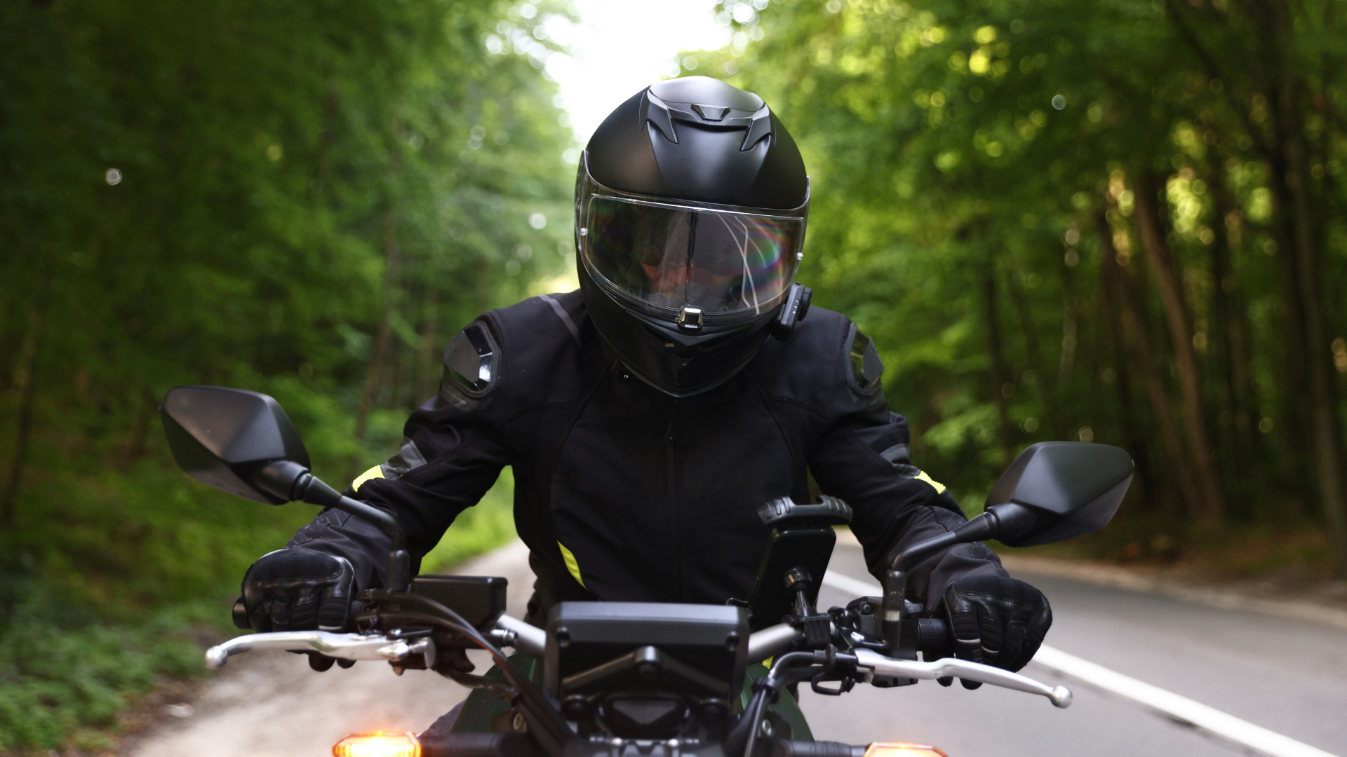 Motorcyclist in black gear riding on a forest road, leaning forward with headlight on