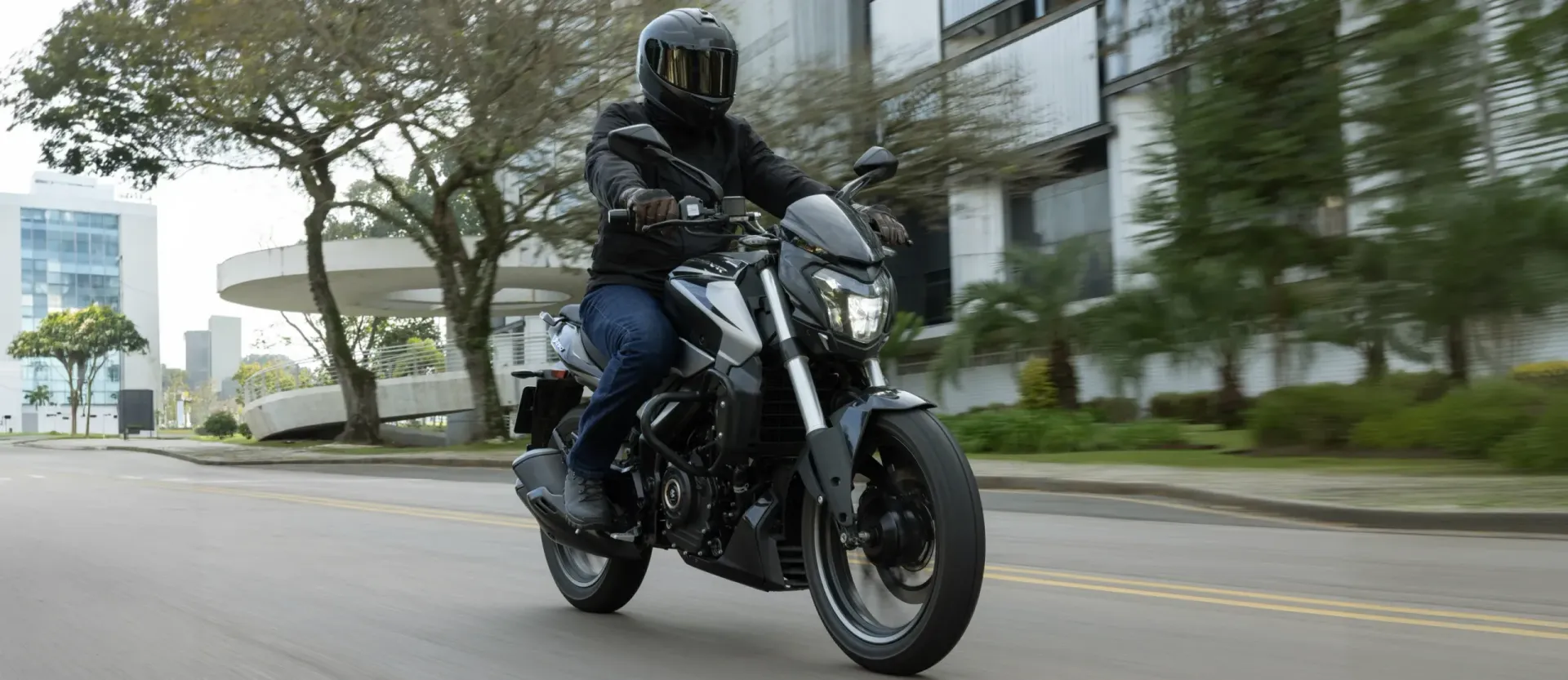 Motorcyclist in black riding a bike down a city street with buildings and trees nearby
