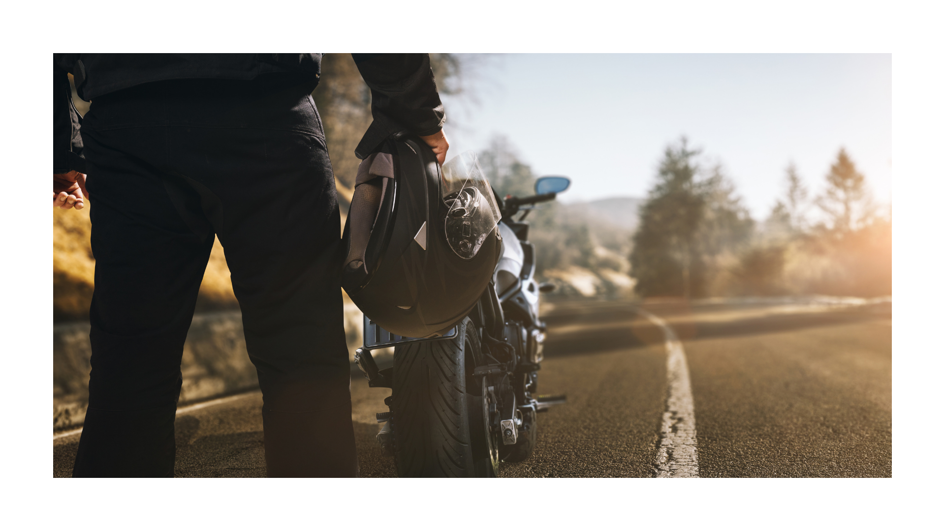 Person stands beside a motorcycle on a sunlit road at sunset