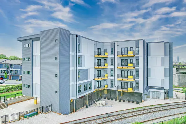 Modern multi-story apartment building, gray and white exterior, with yellow balcony accents, next to train tracks.