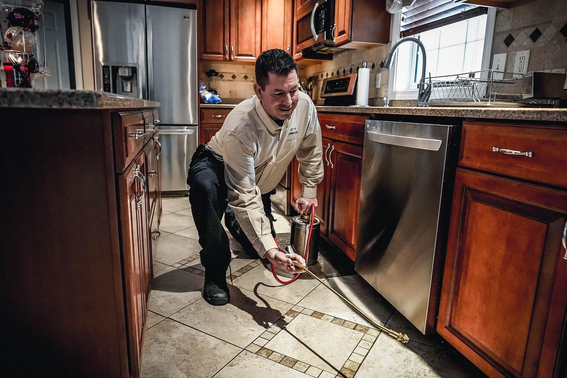 a technician spraying insecticide along kitchen floor tiles near stainless steel dishwasher.