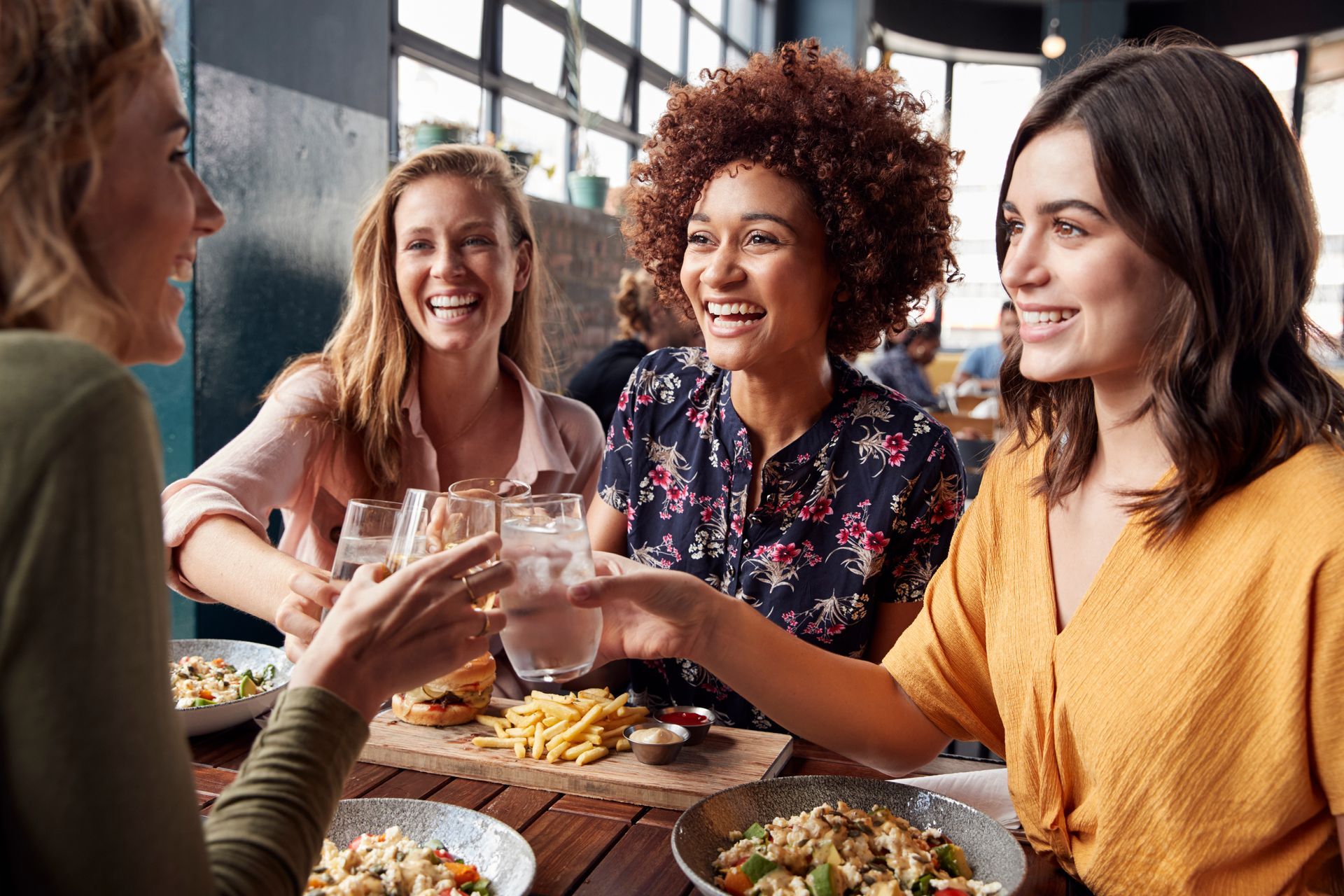 Friends smiling and toasting drinks at a restaurant table with food and window light