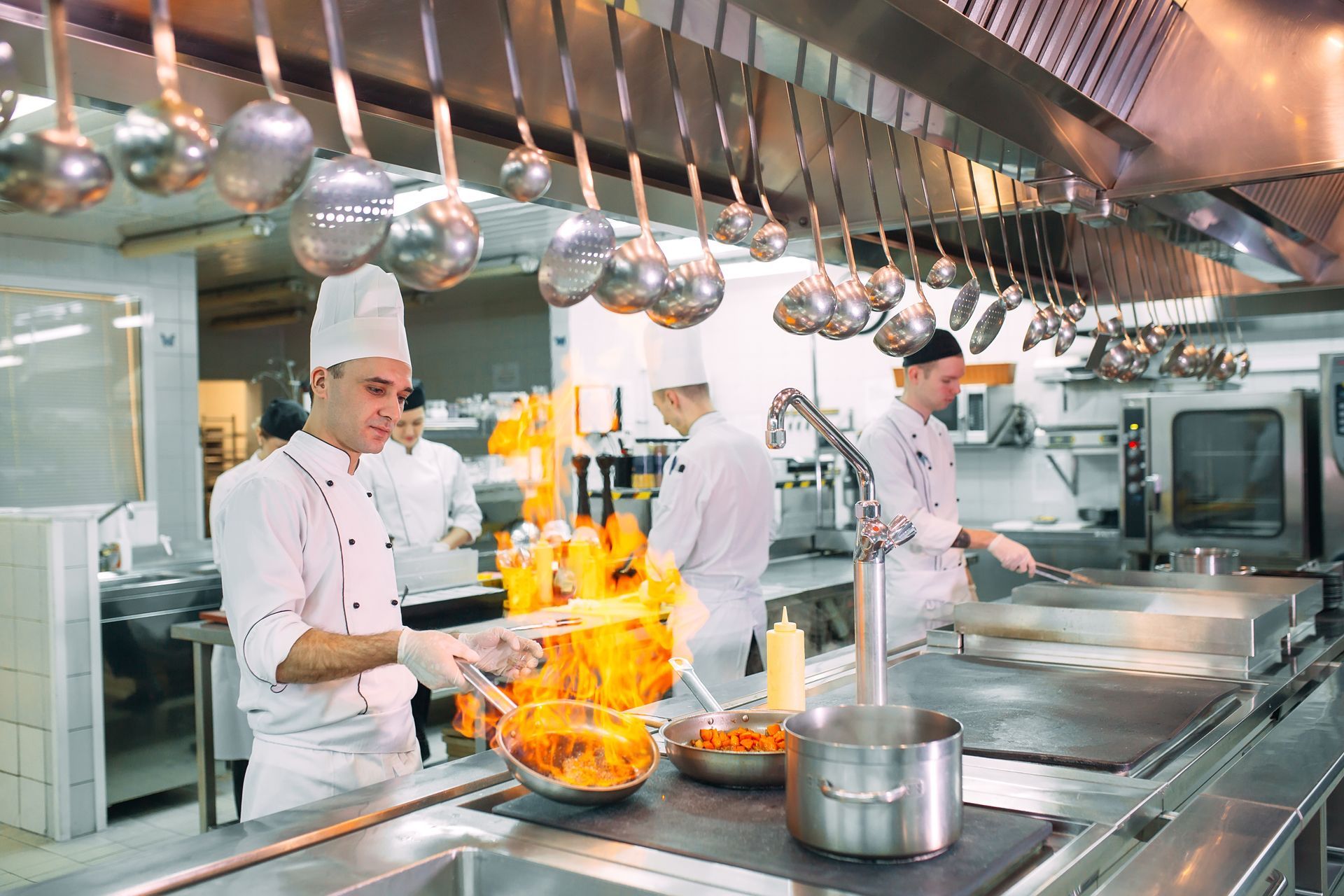 Chefs cooking in a stainless-steel restaurant kitchen, with flames rising from a pan
