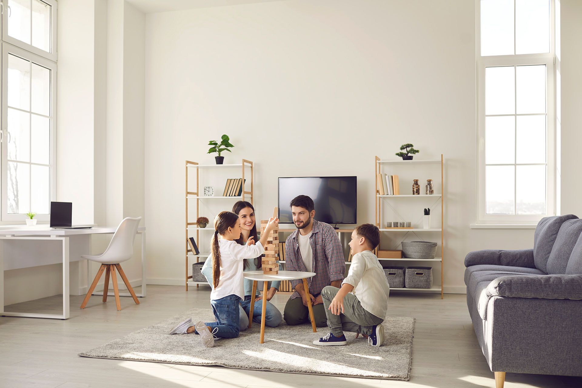 Three people sit on a rug in a bright living room, playing a board game near a TV.