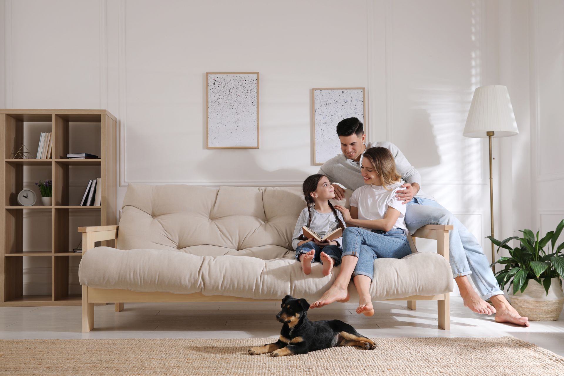 Family sitting on a beige sofa in a bright living room with a small dog on the rug.