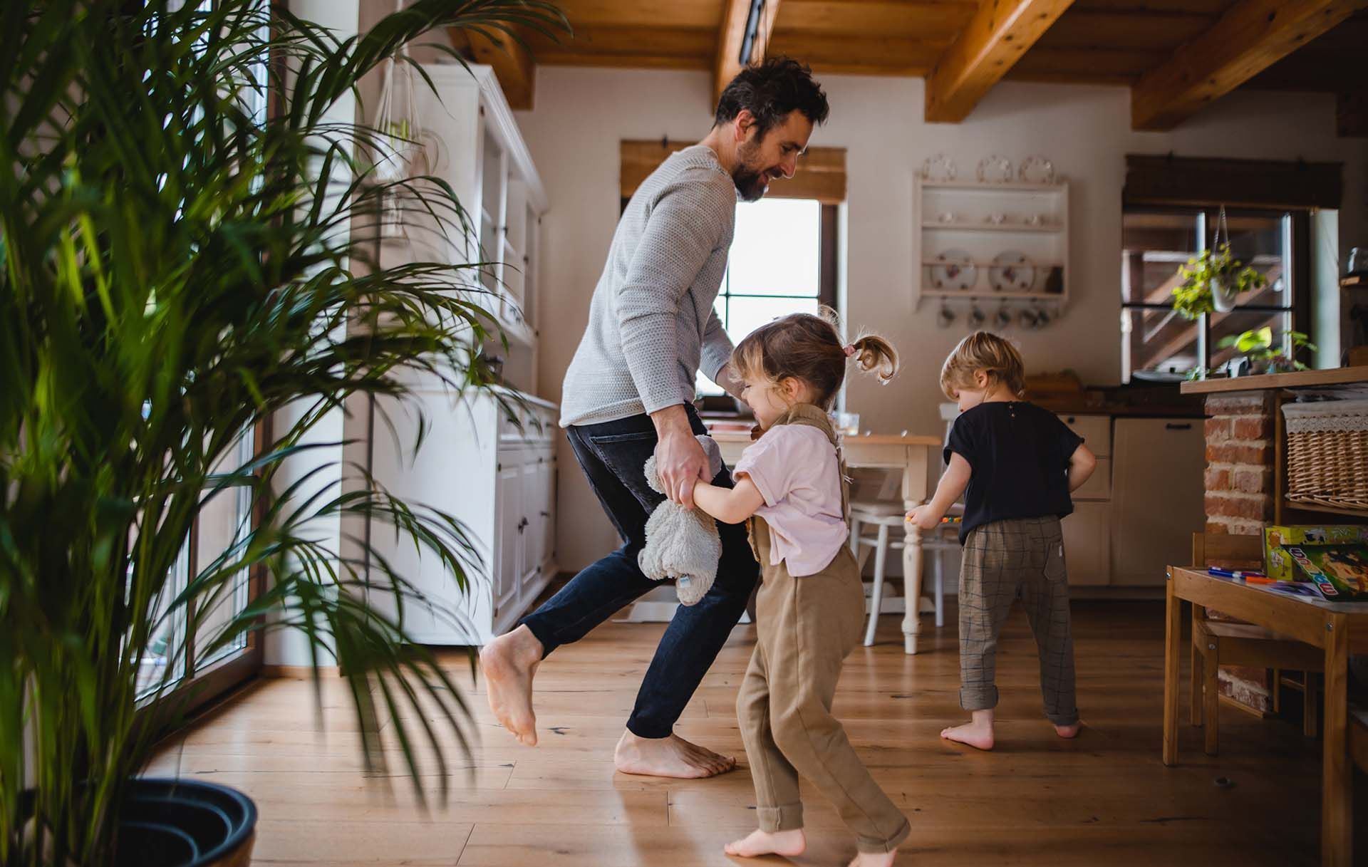 Adult and two children dancing in a bright living room with wooden floors and indoor plants.