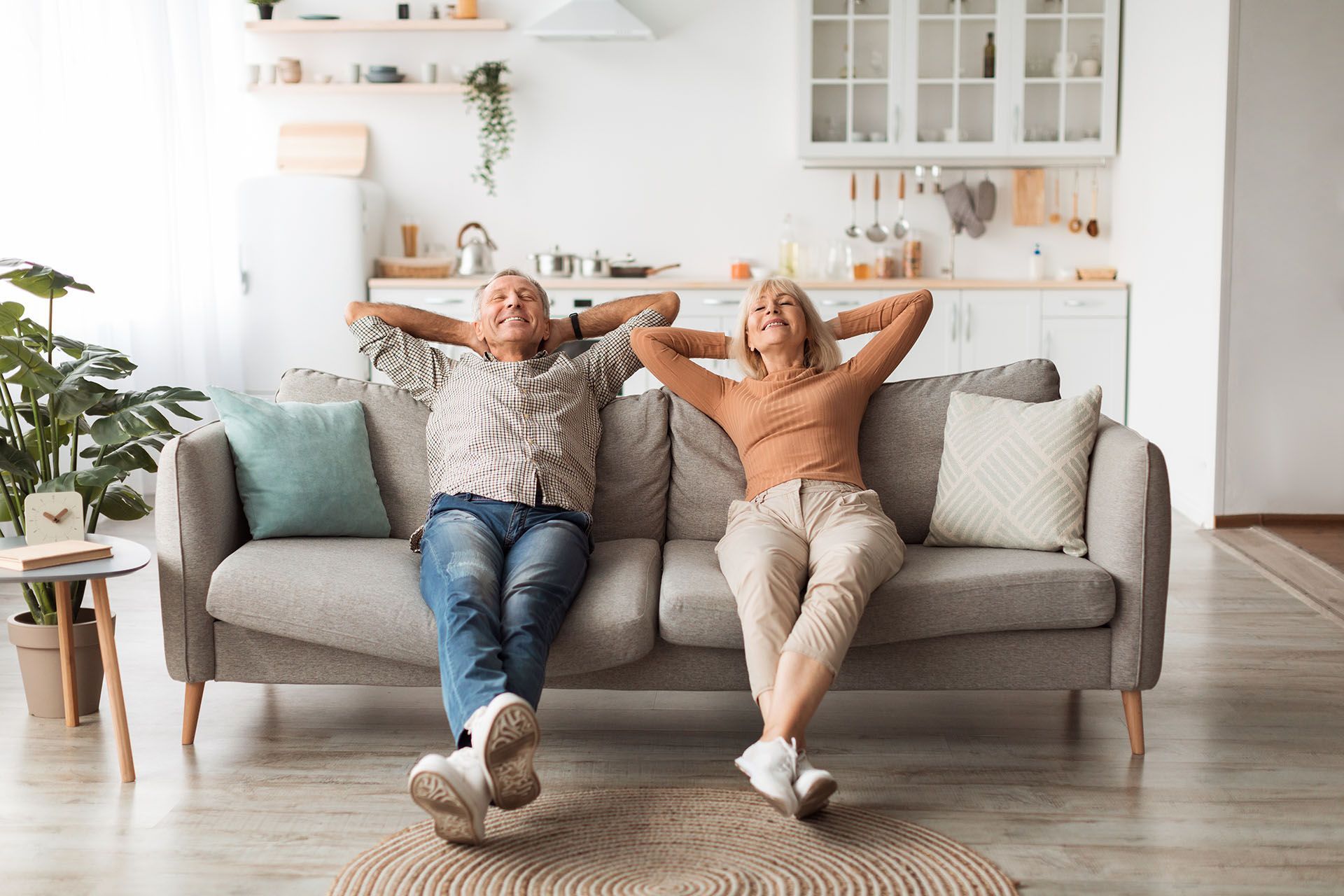 Two people relaxing on a gray sofa in a bright living room, with a plant and shelves in the background. Two people relaxing on a gray sofa in a bright living room, with a plant and shelves in the background.