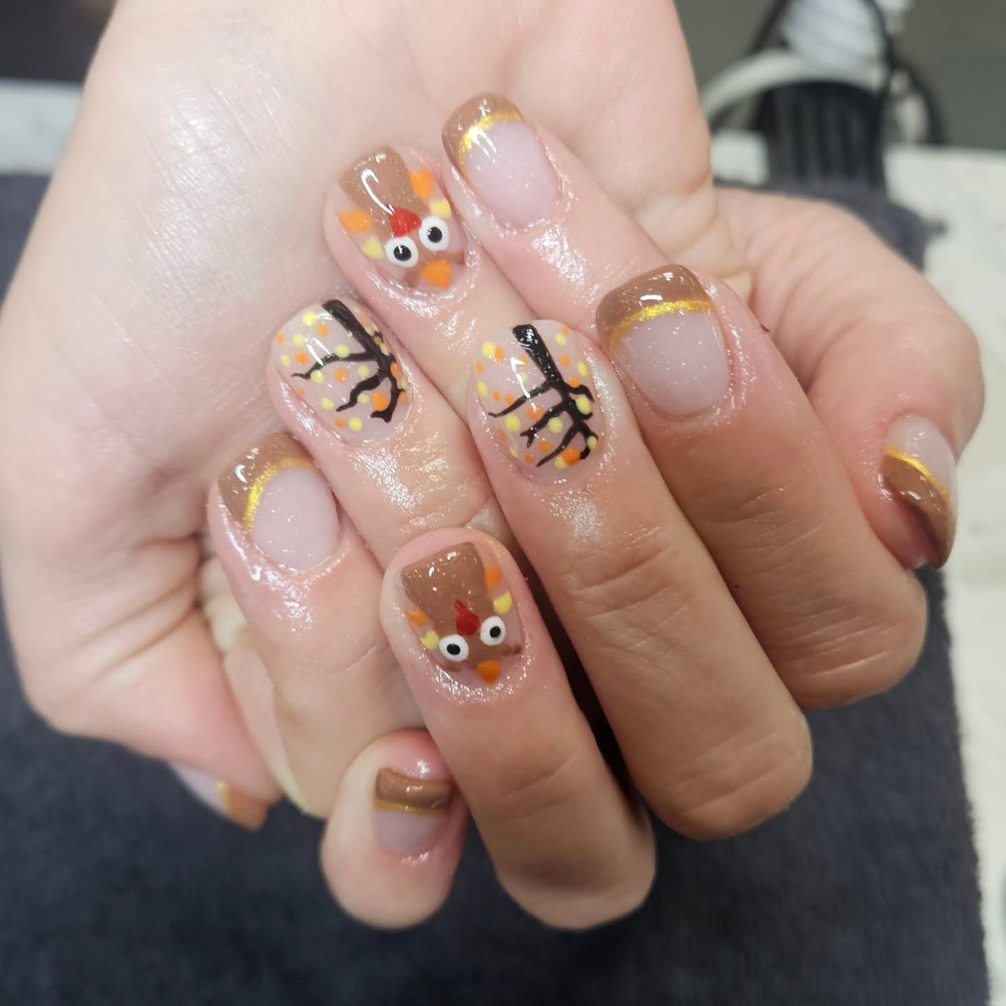A hand features festive Thanksgiving nail art, including turkey designs, autumn branches, and brown-and-gold French tips.
