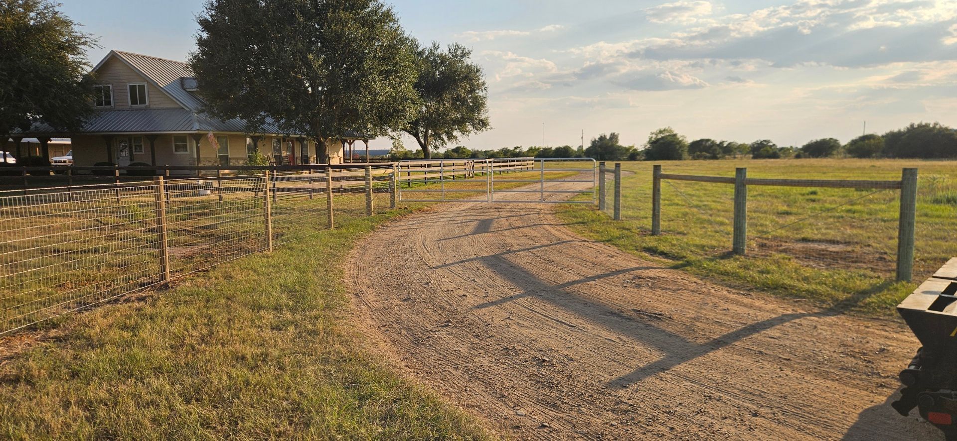 A gravel driveway curves past a house and a wooden fence, with a field in the distance under a cloudy sky.