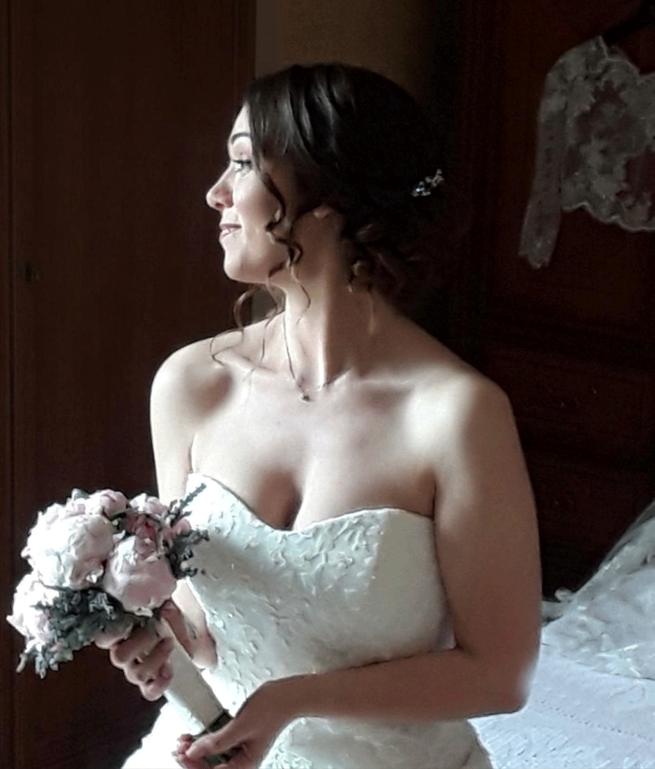 La sposa, in abito bianco senza spalline, tiene in mano un bouquet e guarda di lato in una stanza illuminata da una luce soffusa.