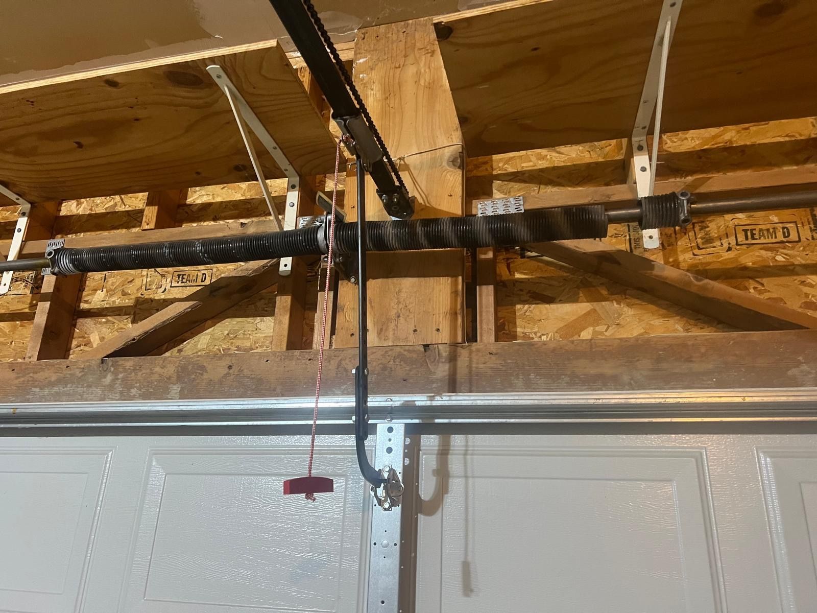 Garage door mechanism with torsion spring, cables, and track mounted on wooden frame.