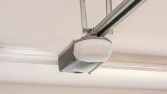Garage door opener unit mounted on the ceiling with green and white casing, visible wiring, and clutter below.
