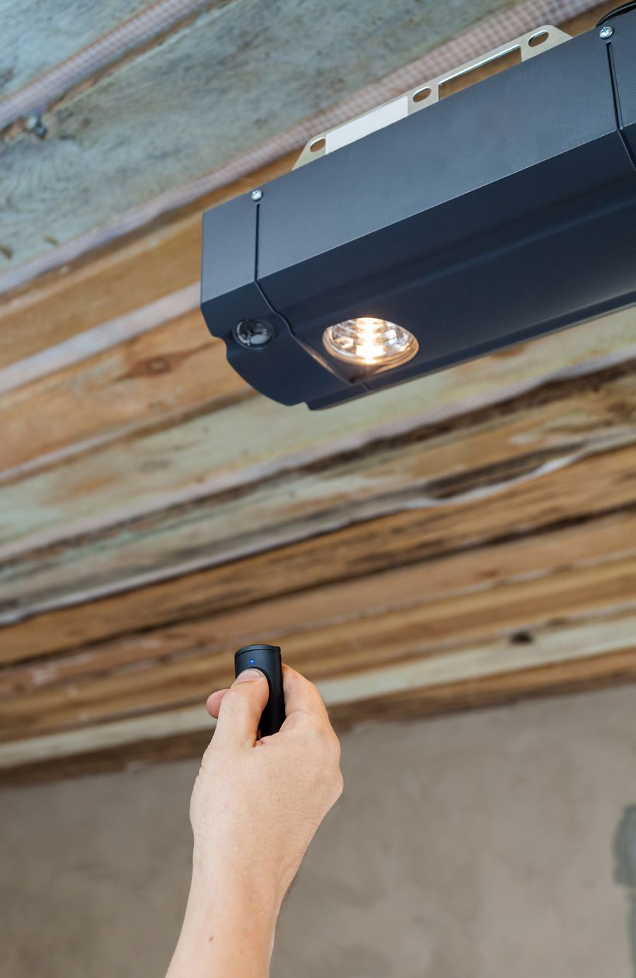 Hand holding remote, aiming it toward a garage door opener with light on, in a wooden ceiling setting.