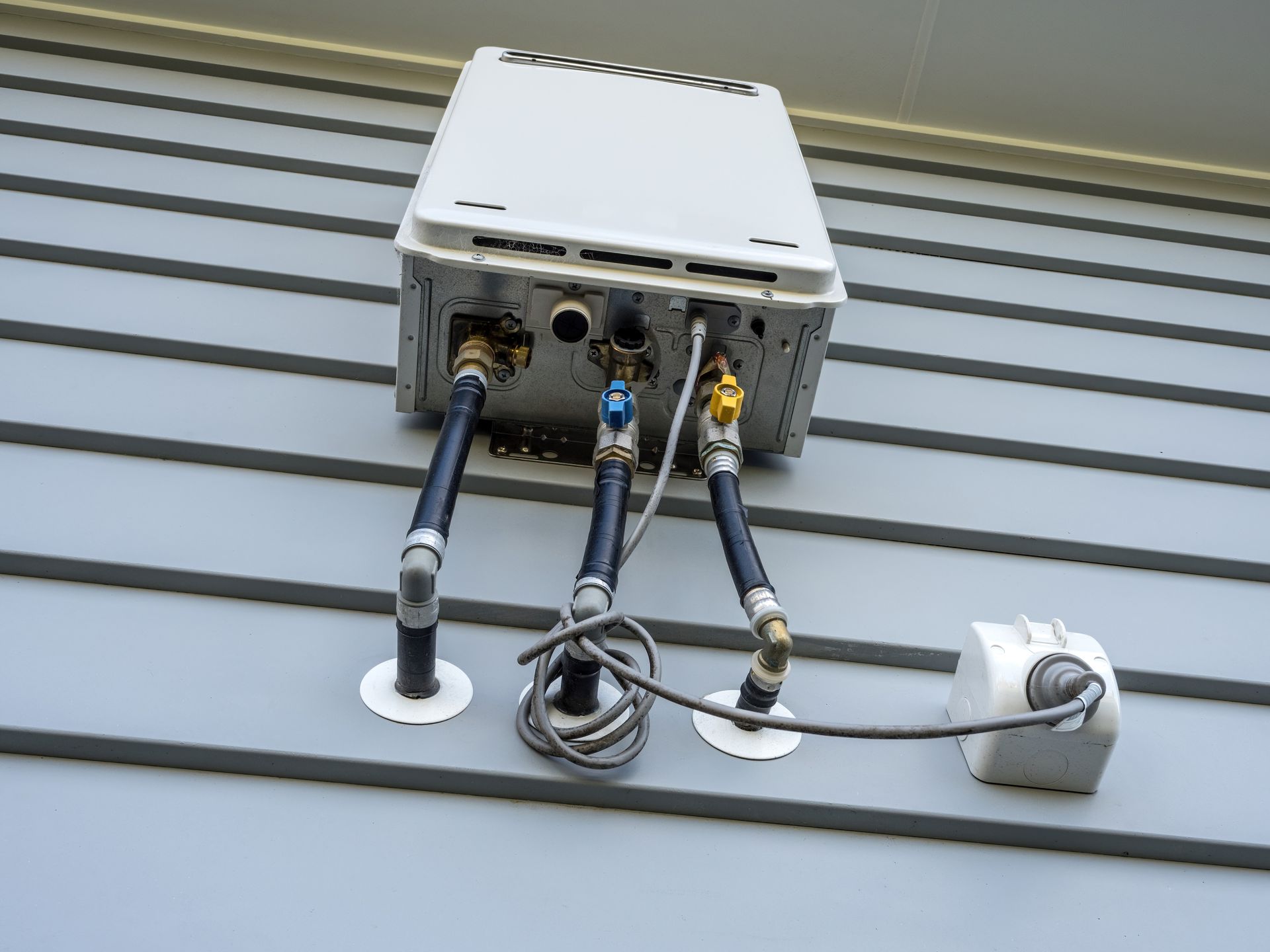 Water heater with pipes and valves mounted on a gray siding wall outdoors