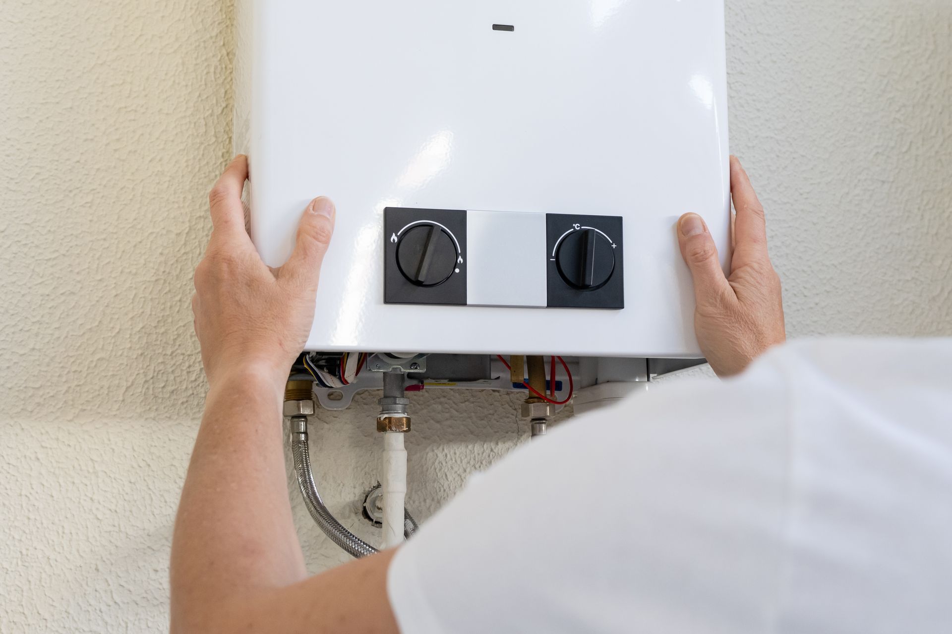 Technician installing a white wall-mounted boiler, adjusting controls and pipes beneath it.