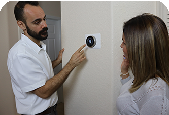 Man points at a thermostat while speaking to a woman. They stand indoors, possibly discussing temperature settings.