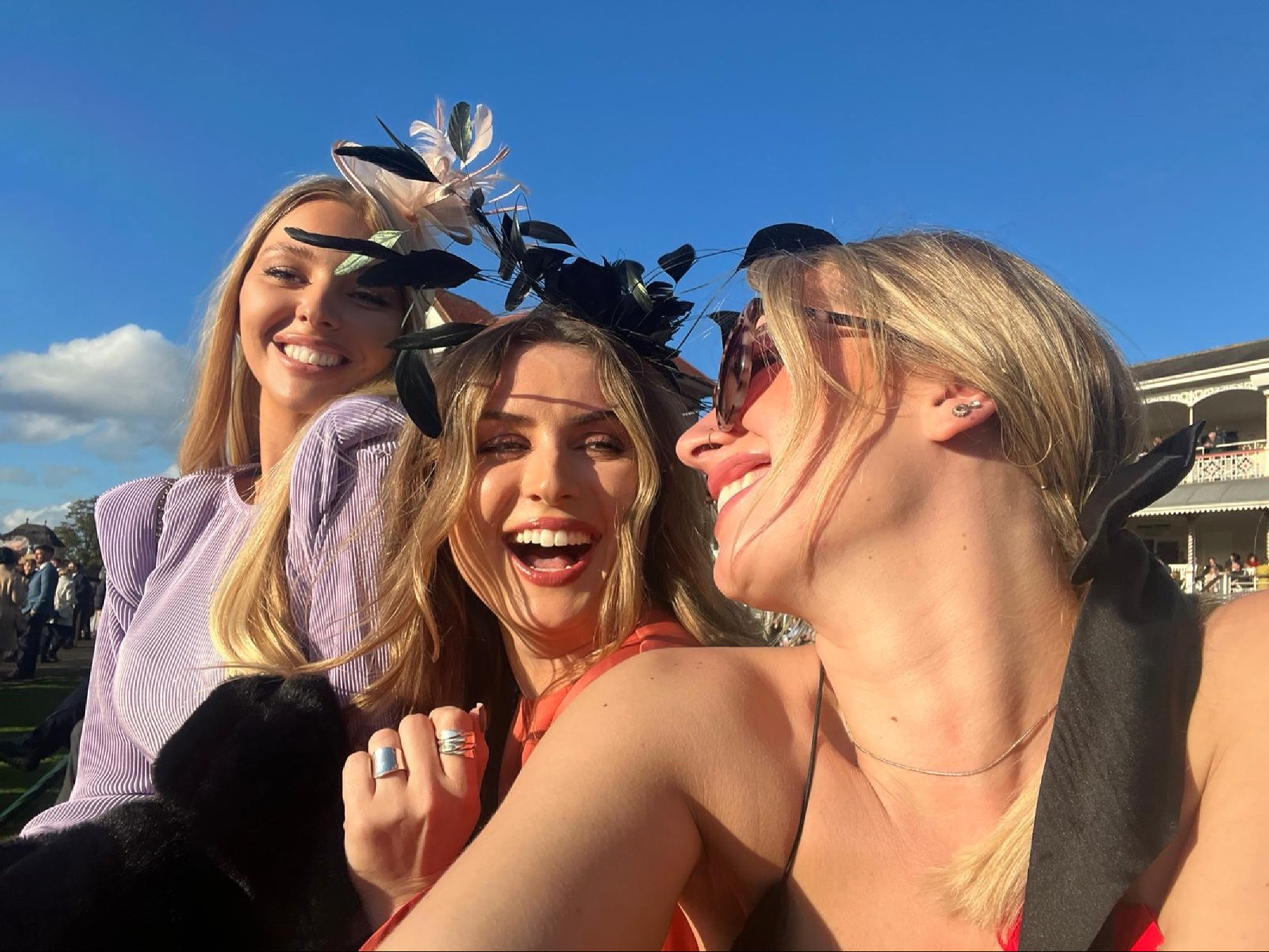 Three women laughing, posing for a selfie at a horse race; one wearing a black headpiece.