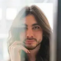 A person with long brown hair and a beard gazes at the viewer while resting their chin on their hand.