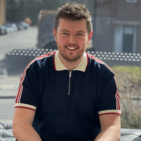 Man smiling, sitting indoors. He has fair skin, wearing a blue polo shirt with red stripes.