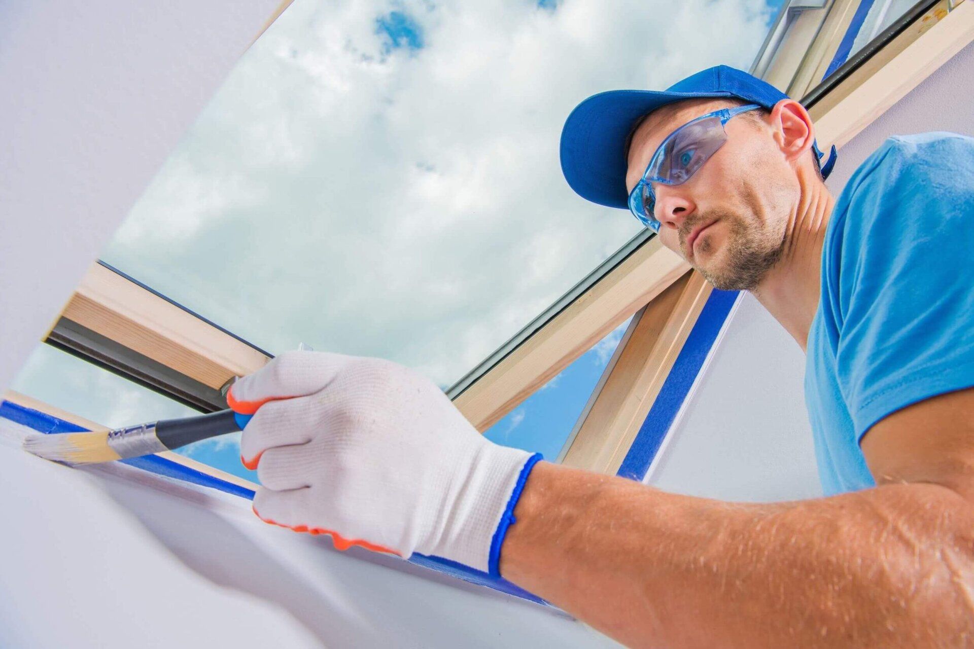 Man in blue shirt and cap paints around a skylight, wearing safety glasses and gloves.