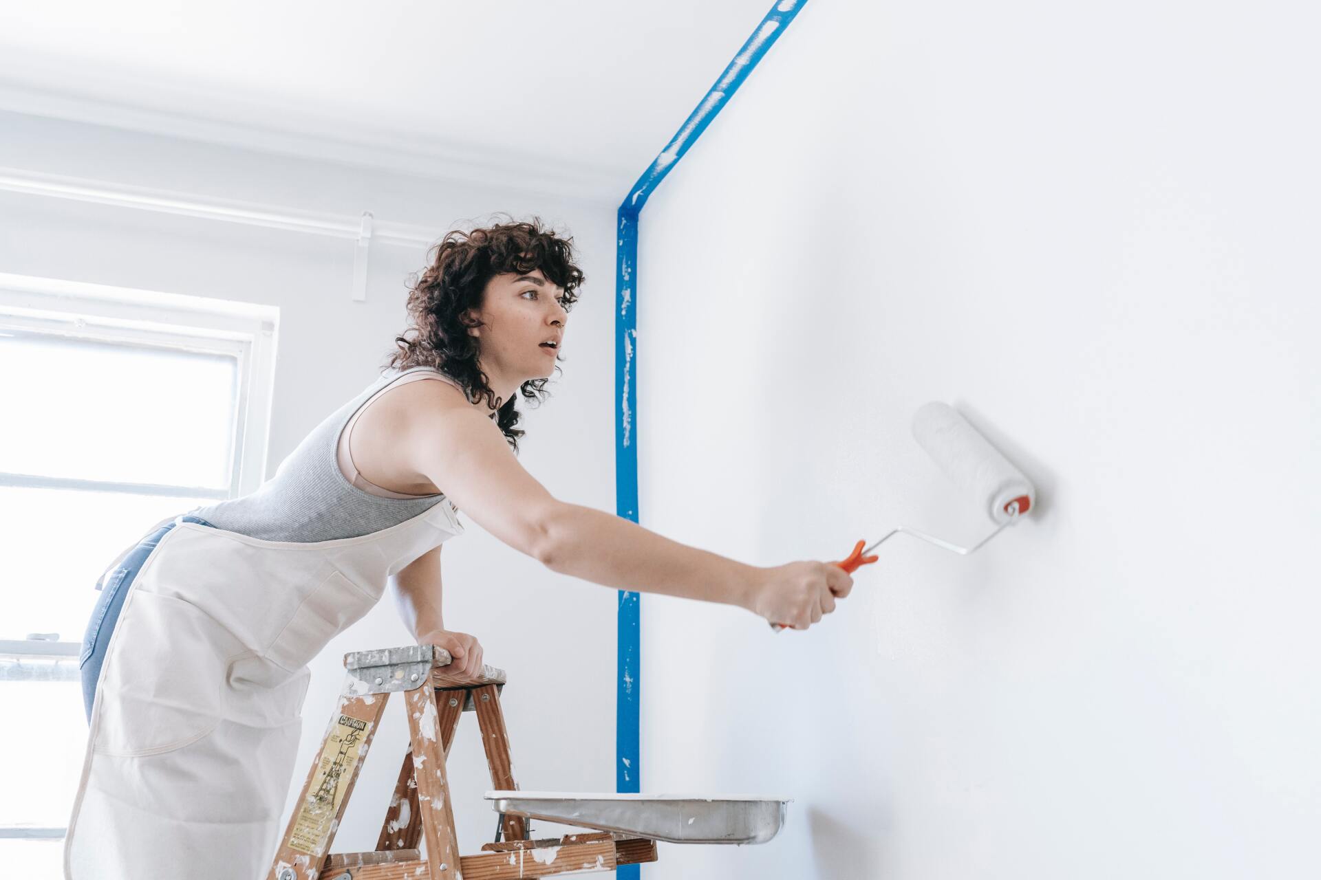 Woman painting a white wall with a roller, standing on a wooden ladder, wearing an apron.