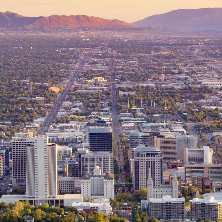 View of Salt Lake City, Utah, with downtown buildings, streets, and mountains in the background under a warm, golden sky. View of Salt Lake City, Utah, with downtown buildings, streets, and mountains in the background under a warm, golden sky.