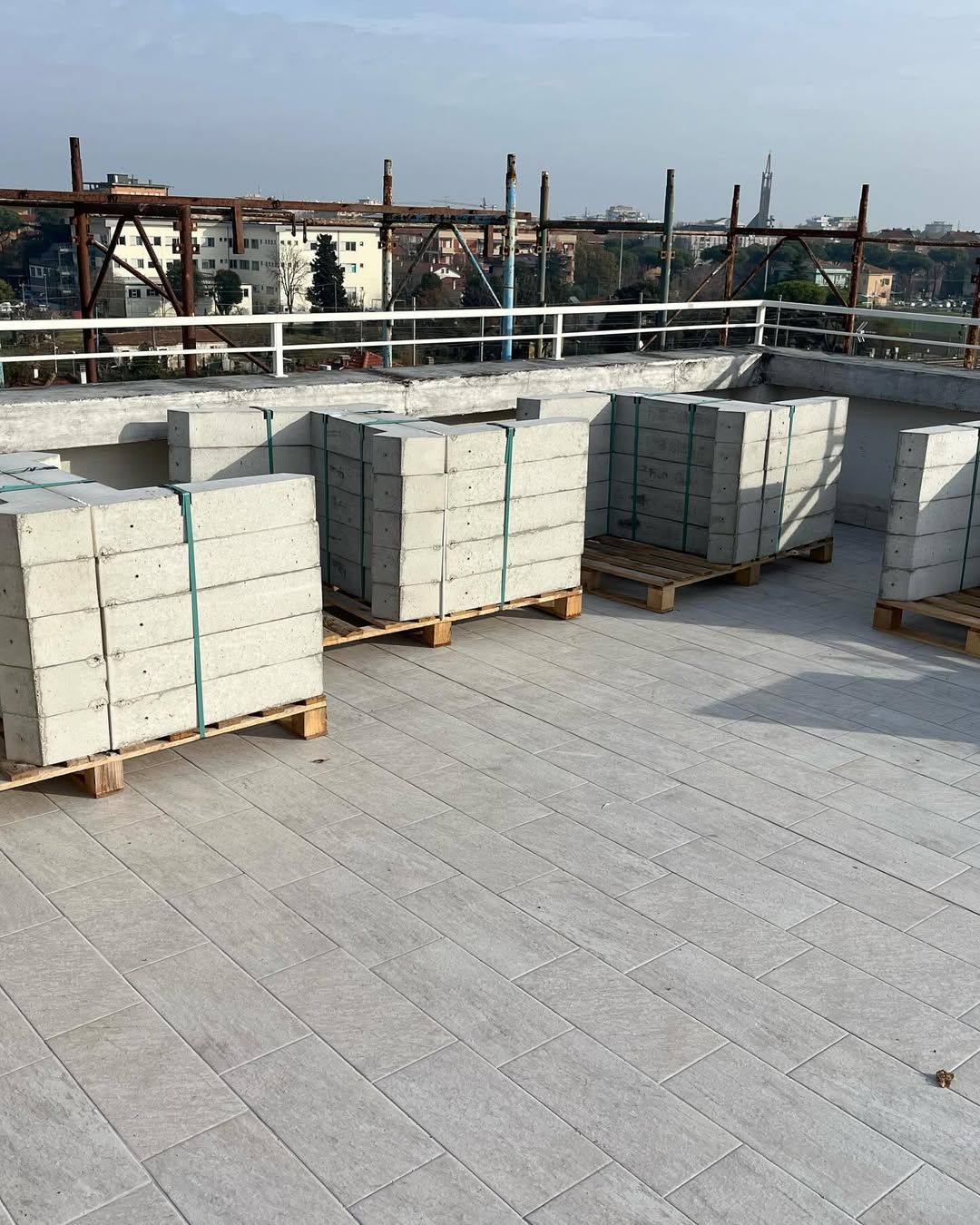 Quattro bancali di blocchi di cemento grigio impilati sono posizionati su una terrazza pavimentata sul tetto, con vista sulla città sullo sfondo.