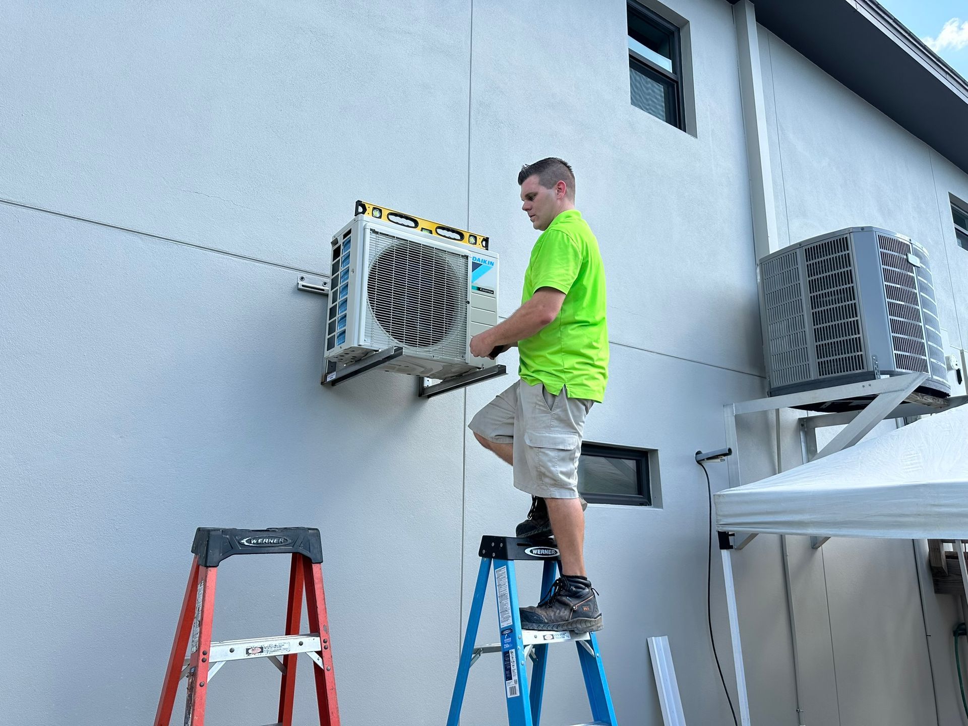 Worker on ladder servicing an outdoor air conditioner unit beside a building